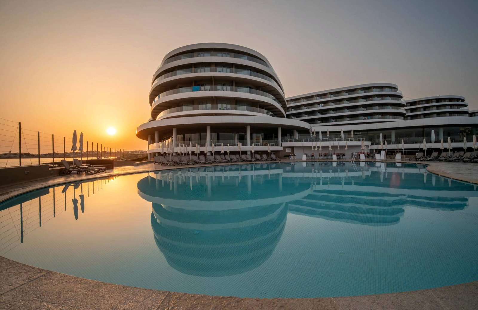 The curved, tiered blocks of Ramla Bay Resort behind a large outdoor pool, with rows of folded parasols along the terrace and the sun setting over the sea to the left.