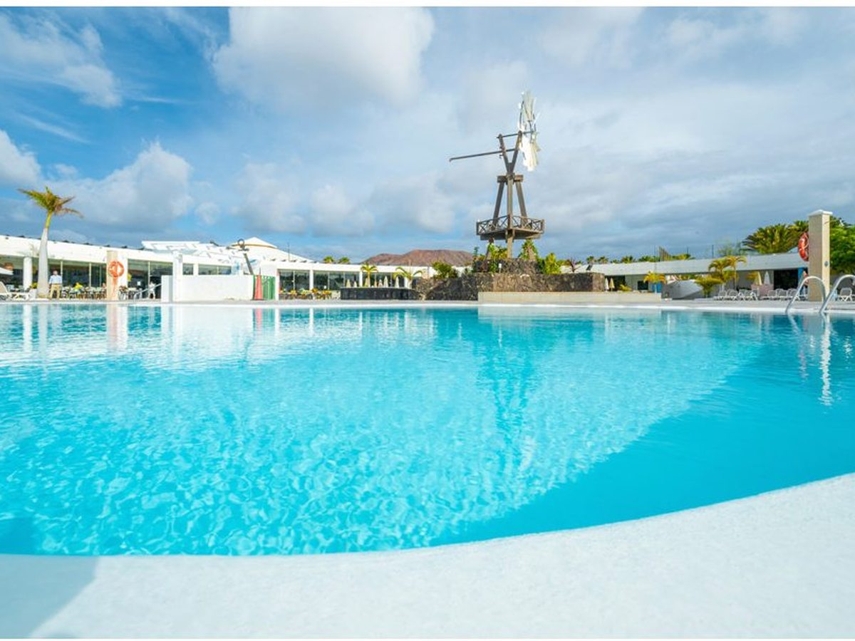 A large outdoor swimming pool with clear turquoise water, a wooden windmill on a rock feature behind it and low white resort buildings and palms beyond.