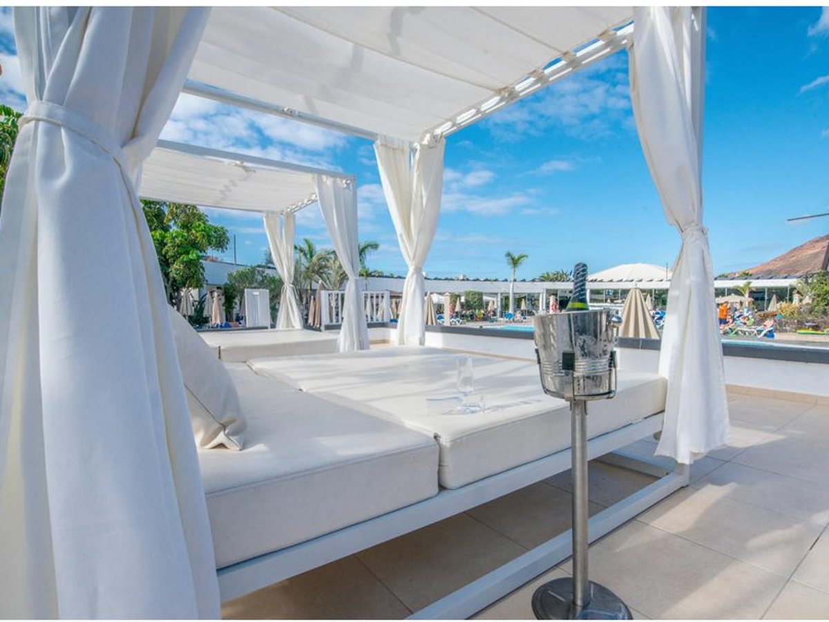 A white curtained daybed with a thick mattress on a tiled poolside terrace, an ice bucket and glasses beside it, with parasols, sun loungers and the pool behind.