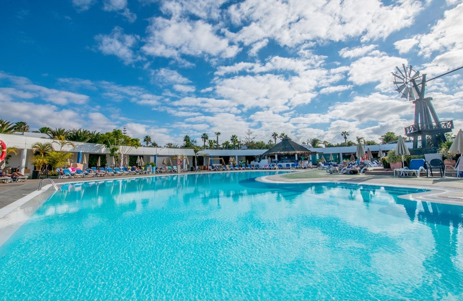 A large outdoor pool with turquoise water at Relaxia Lanzasur Club, edged with sun loungers and a thatched pool bar, with single-storey white bungalow buildings, palms and a windmill behind.