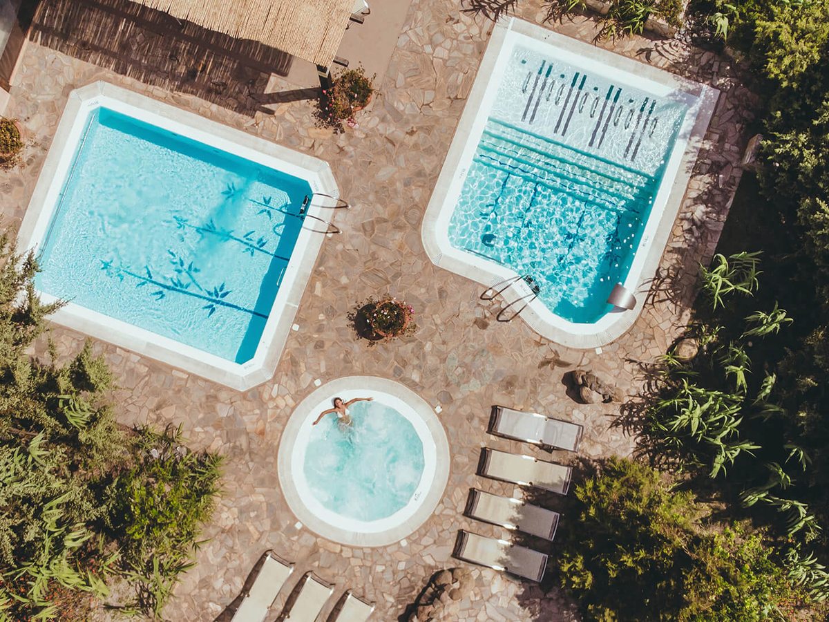 Aerial view of two rectangular spa pools and a round bubbling jacuzzi set in a stone-paved terrace among green shrubs, with sun loungers alongside.