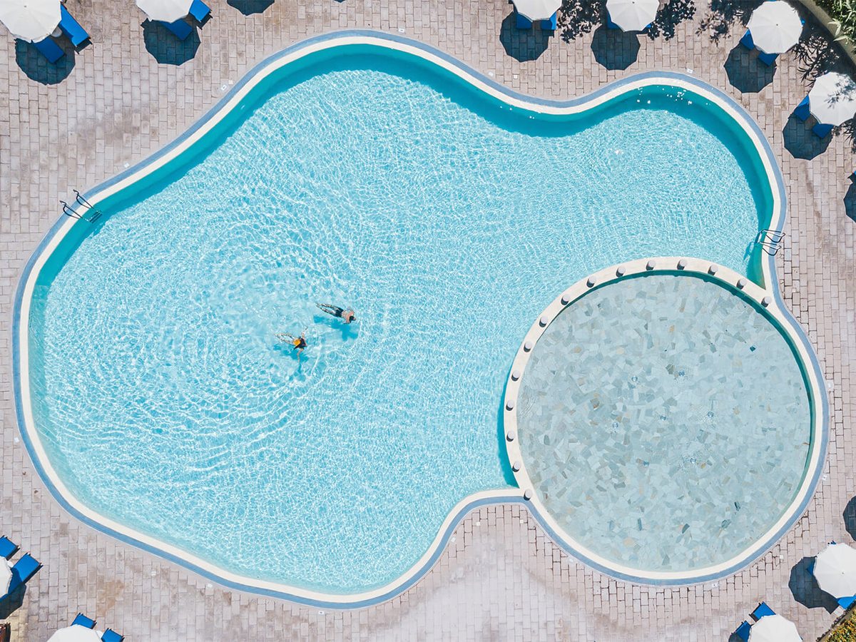Aerial view of a large freeform pool with a small round shallow pool joined to one end, ringed by white parasols and sun loungers on pink paving.