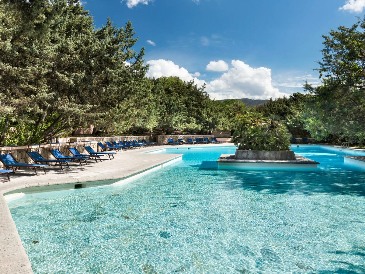 A wide pool with a raised planted island in the middle, blue sun loungers along the paved edge and juniper trees behind.