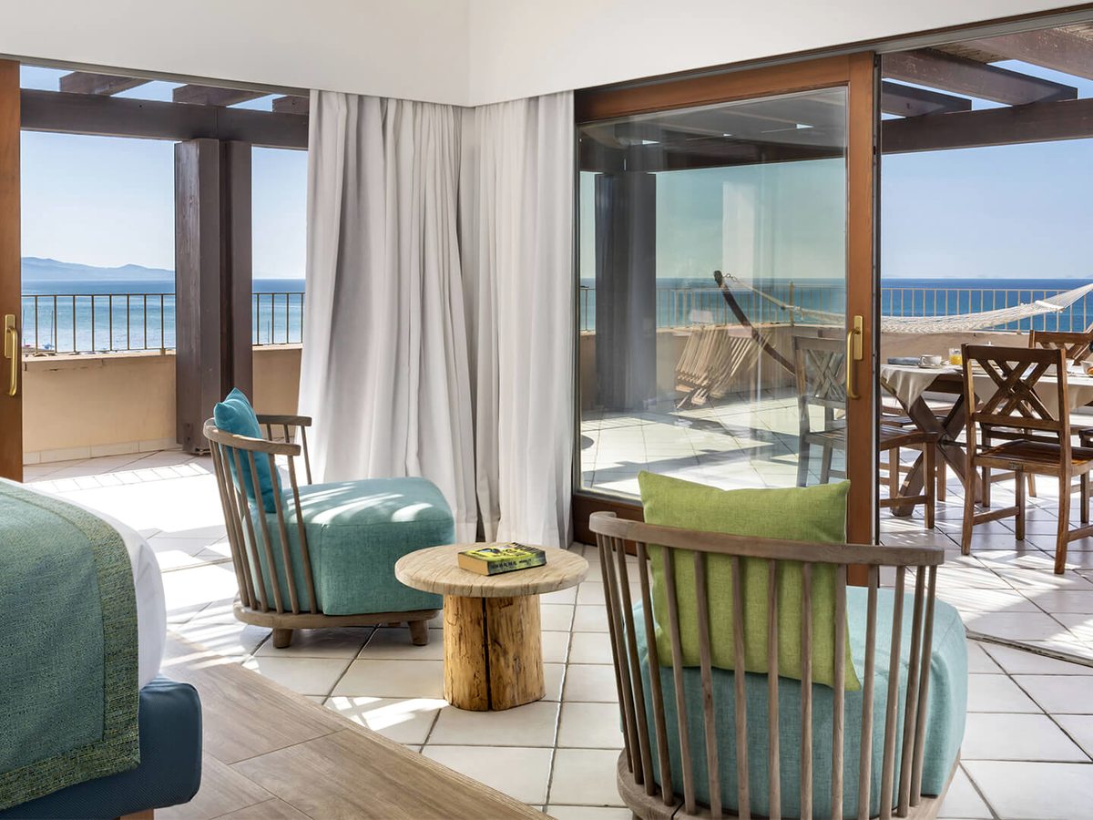 Corner of a guest room with green and blue armchairs and a low wooden table, and sliding doors onto a tiled terrace with a hammock and a dining table above the sea.