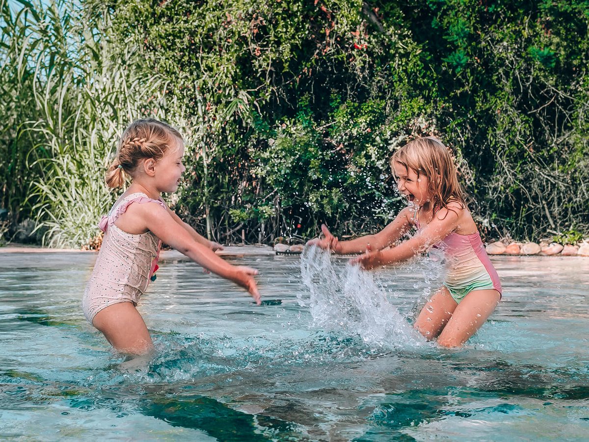 Two young girls in swimsuits splashing each other in shallow pool water, with tall grasses and shrubs behind them.