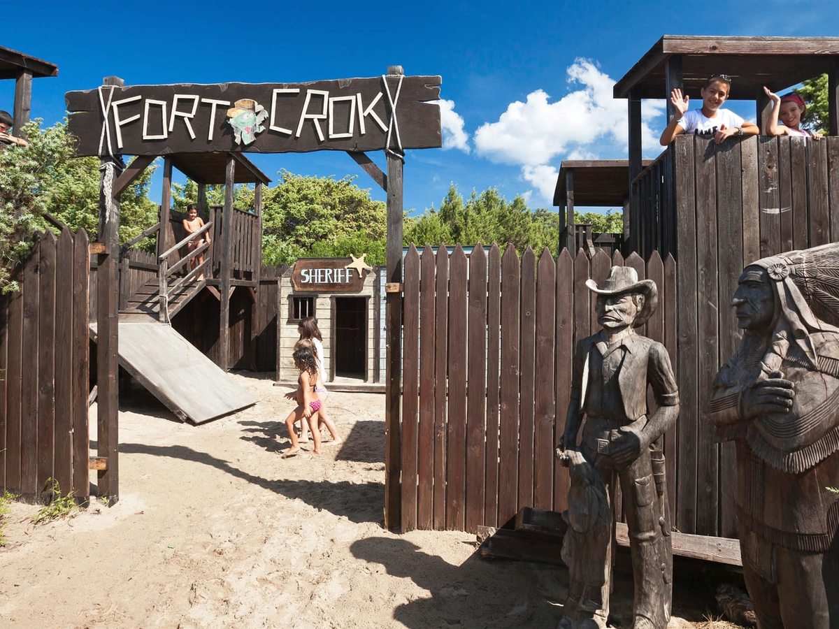 A wooden cowboy fort with a Fort Crok sign, a sheriff's hut, a slide and carved wooden figures, with children playing on the sand around it.