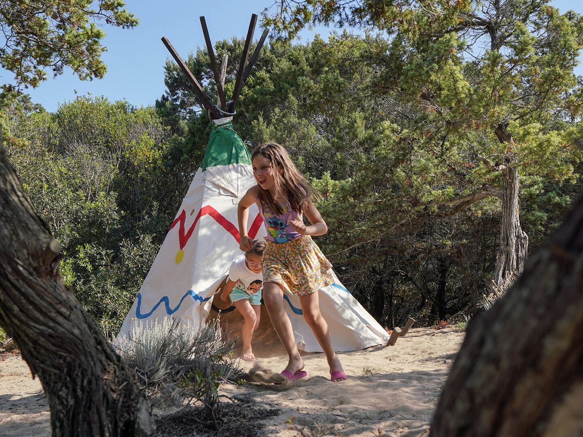 A painted teepee among juniper bushes on a sandy path, with a girl running out of it towards the camera and a younger child crouching in the entrance.