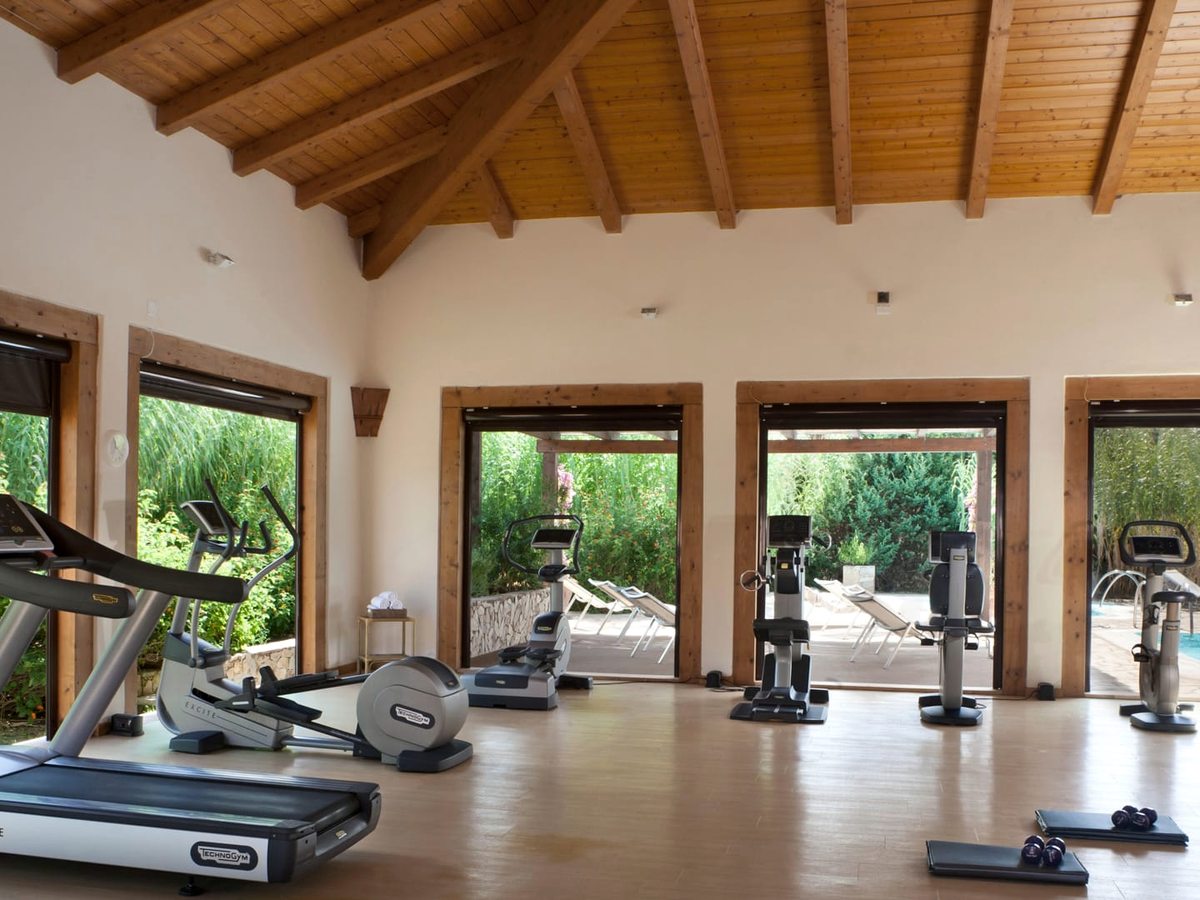 A gym with treadmills, cross-trainers and exercise bikes under a beamed wooden ceiling, with dumbbells and mats on the floor and open doors onto greenery.