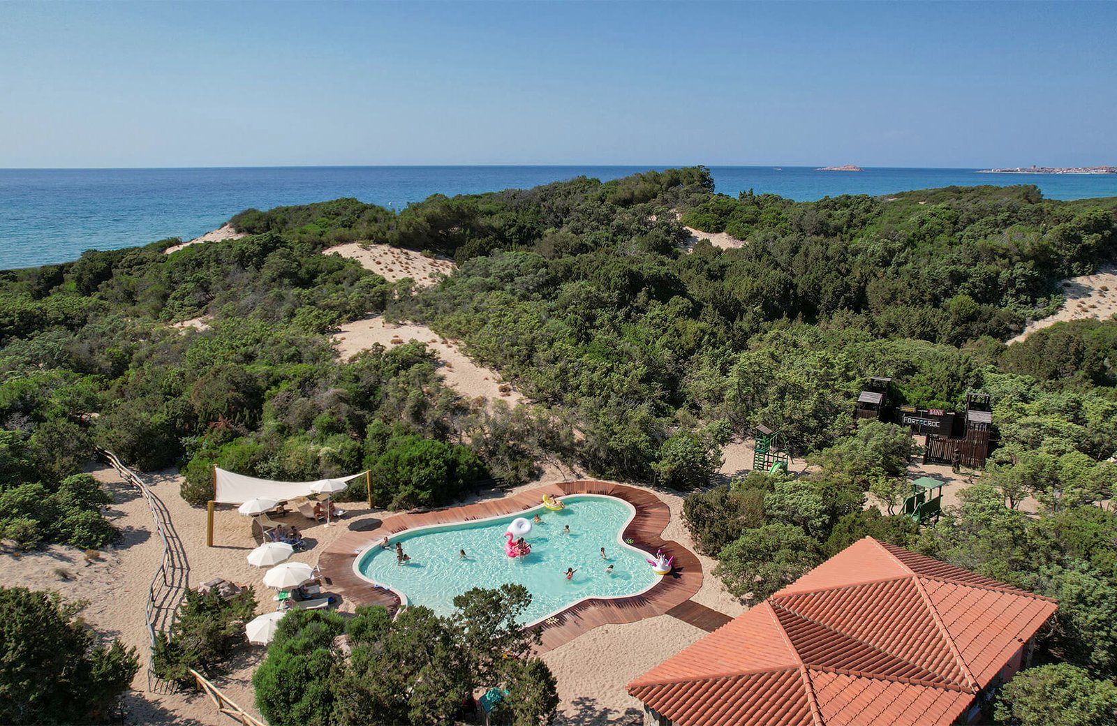Aerial view of a freeform children's pool with a timber surround and shade sails, set among sand dunes and juniper scrub, with a wooden cowboy fort to one side and the sea behind.