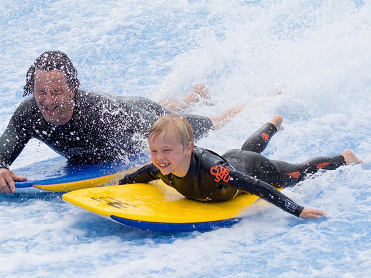 A child lying on a yellow bodyboard riding the FlowRider's sheet of white water, with an instructor in a wetsuit alongside.