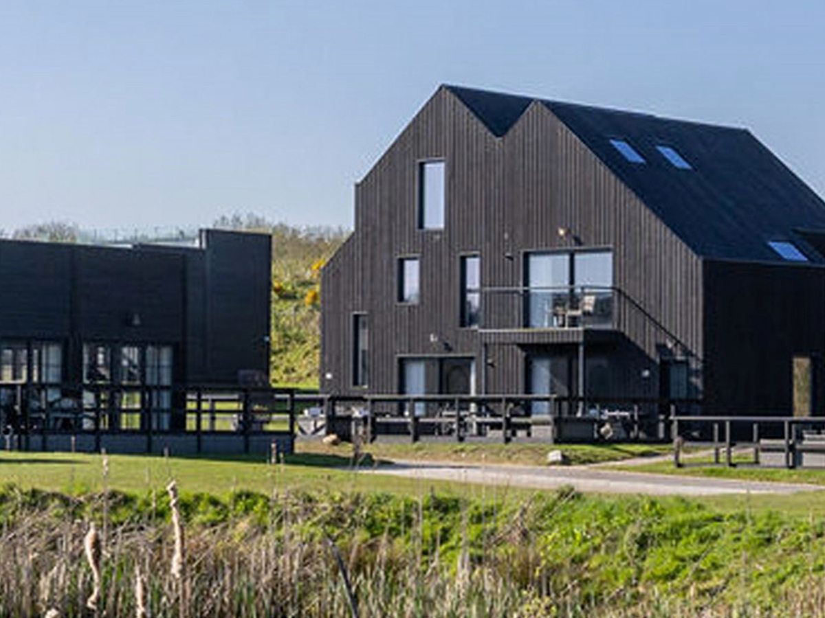 Tall timber-clad barns with slate-dark roofs and first-floor balconies standing on a grassy slope above a reed-fringed lake.