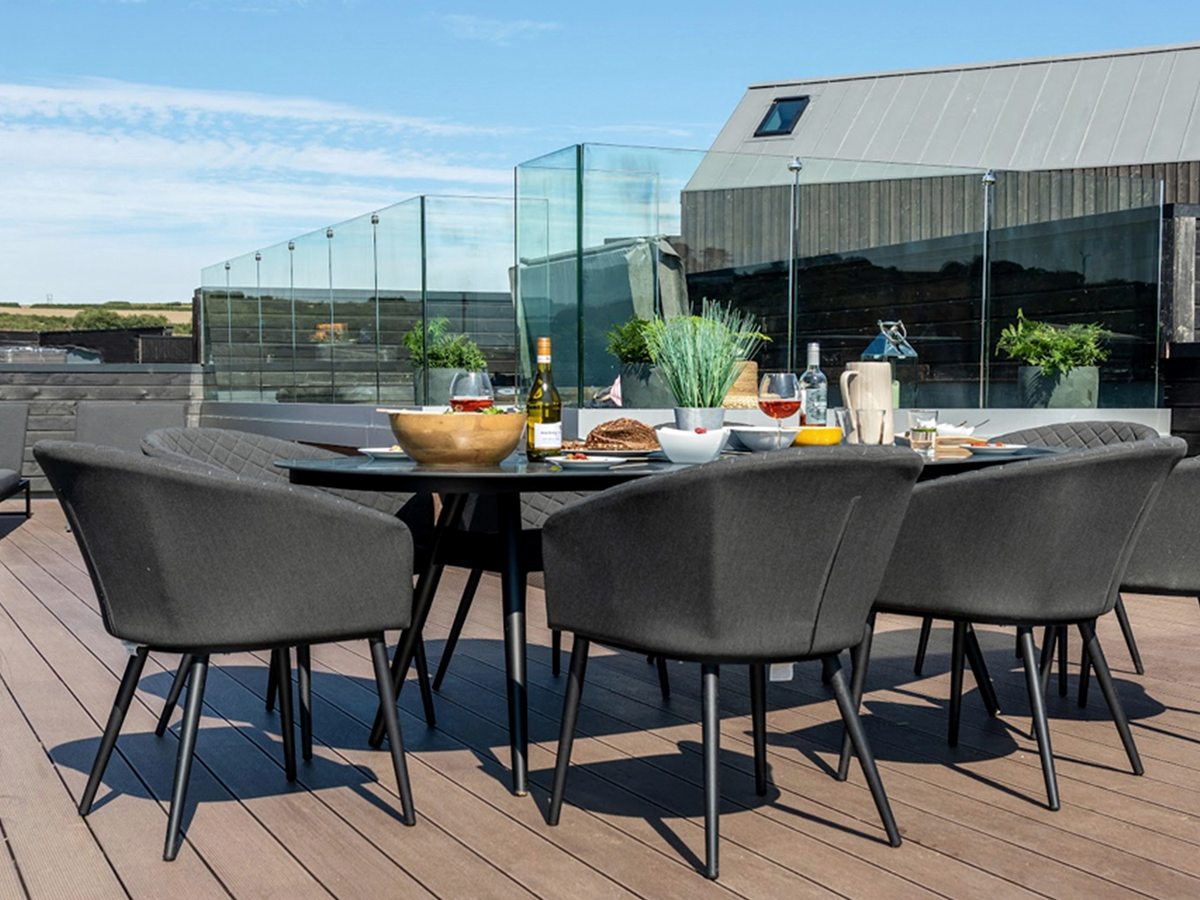 A round outdoor dining table set with food and wine, surrounded by grey upholstered chairs on a composite-decked roof terrace with a glass balustrade and open countryside beyond.