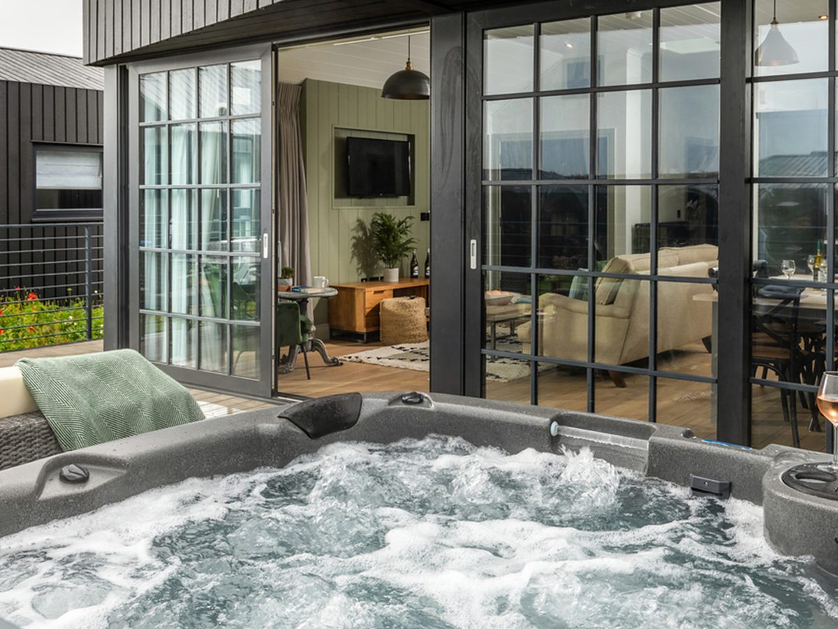 A bubbling hot tub on a decked terrace outside a lodge, with folding glass doors open onto the lounge and dining area behind.