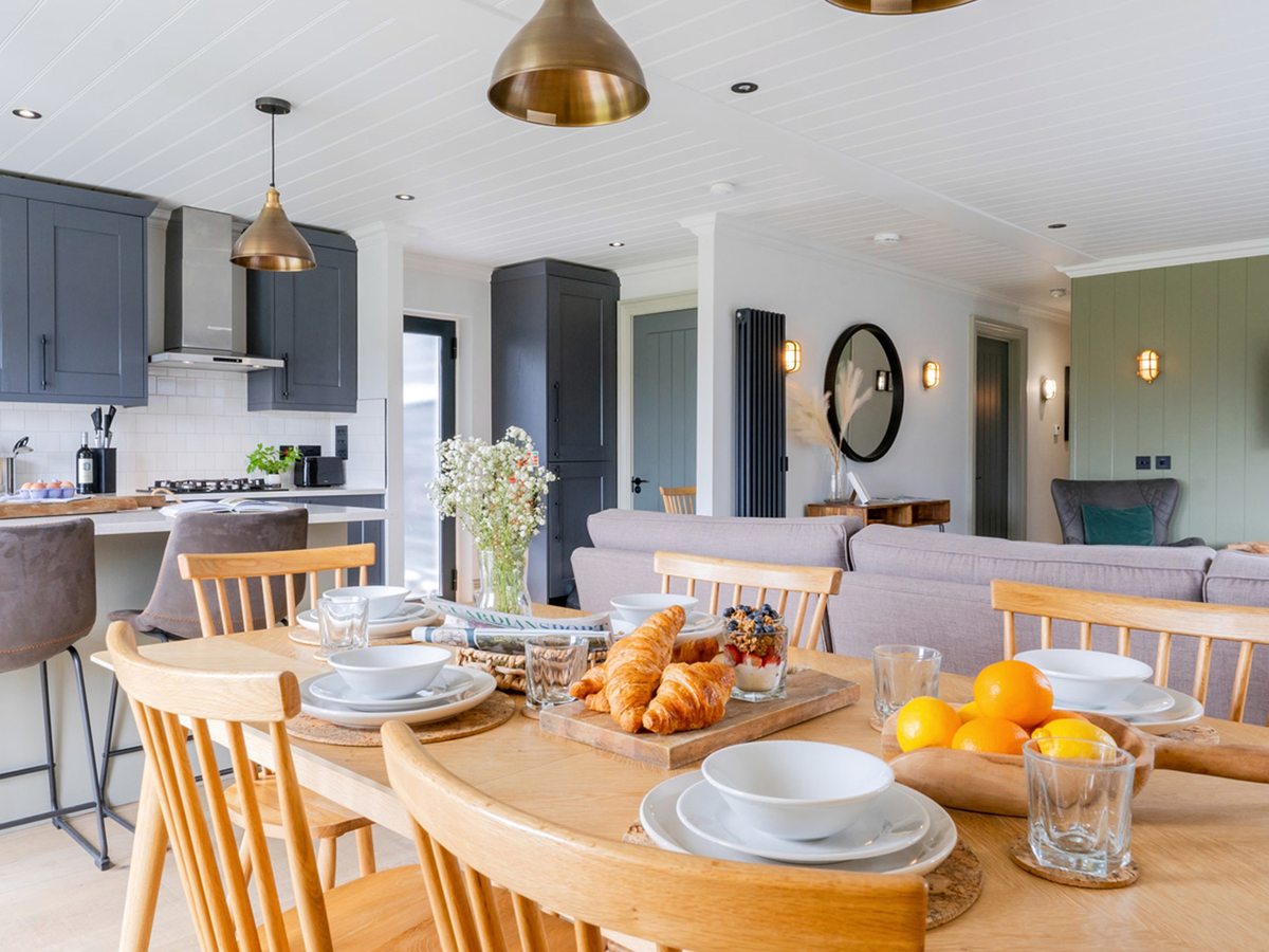 An oak dining table laid for breakfast with croissants, fruit and bowls, in front of a navy shaker kitchen with a breakfast bar and brass pendant lights, opening through to a lounge.