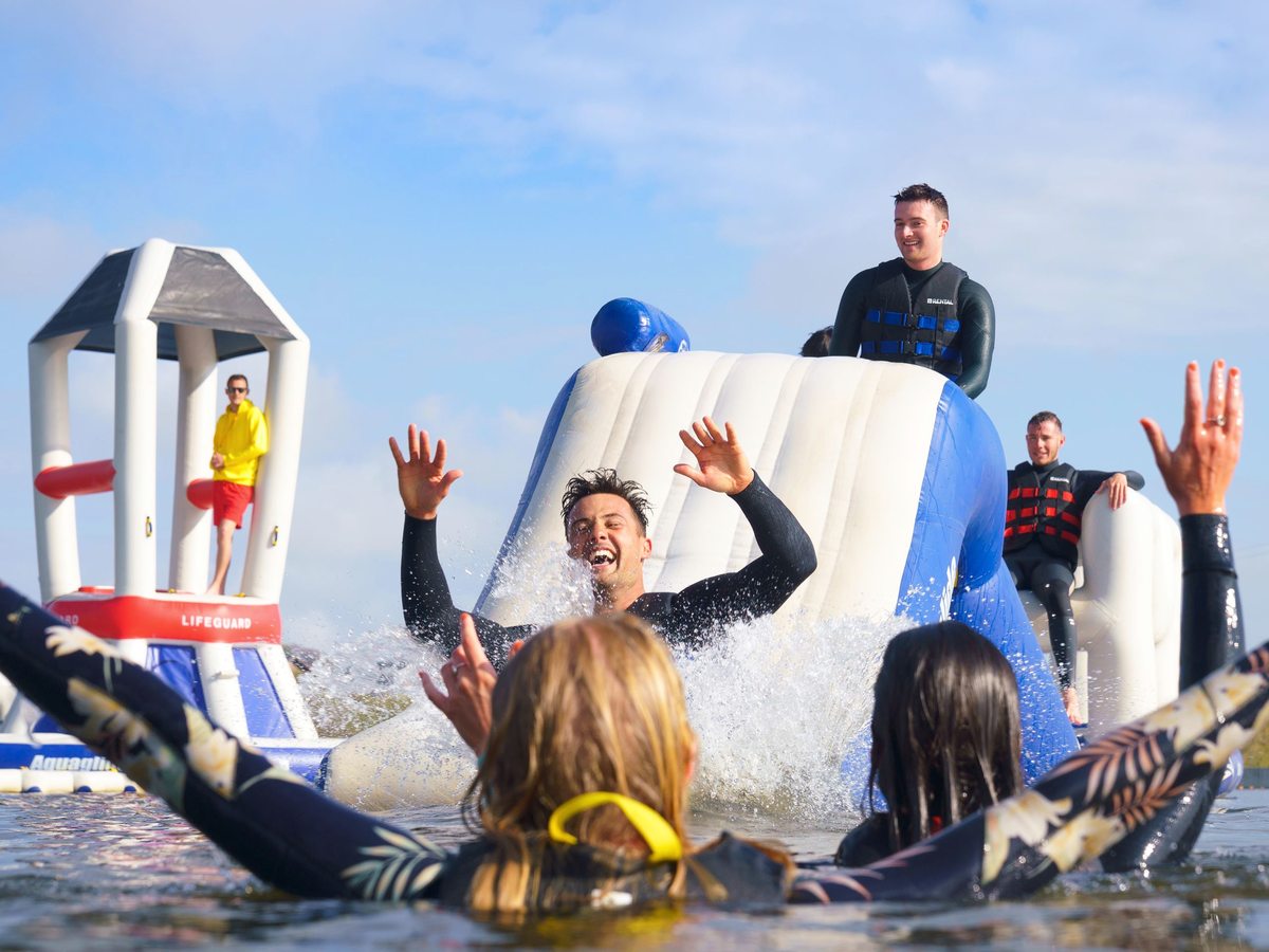 Adults in wetsuits and buoyancy aids falling into the water off a large blue and white inflatable course on the lake, with a lifeguard on the platform behind.