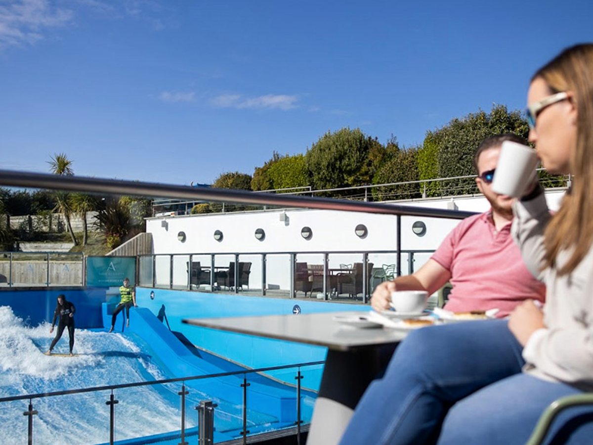 Two people drinking hot drinks at an outdoor table, with the blue FlowRider surf pool and its white timber-clad building behind them.