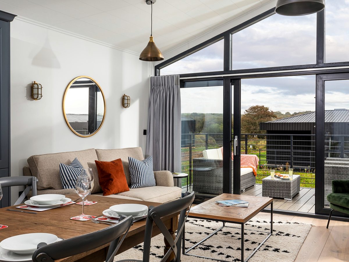 A lodge interior with a wooden dining table laid for a meal, a cream sofa with striped and rust cushions, and full-height glass doors onto a decked balcony with rattan seating.