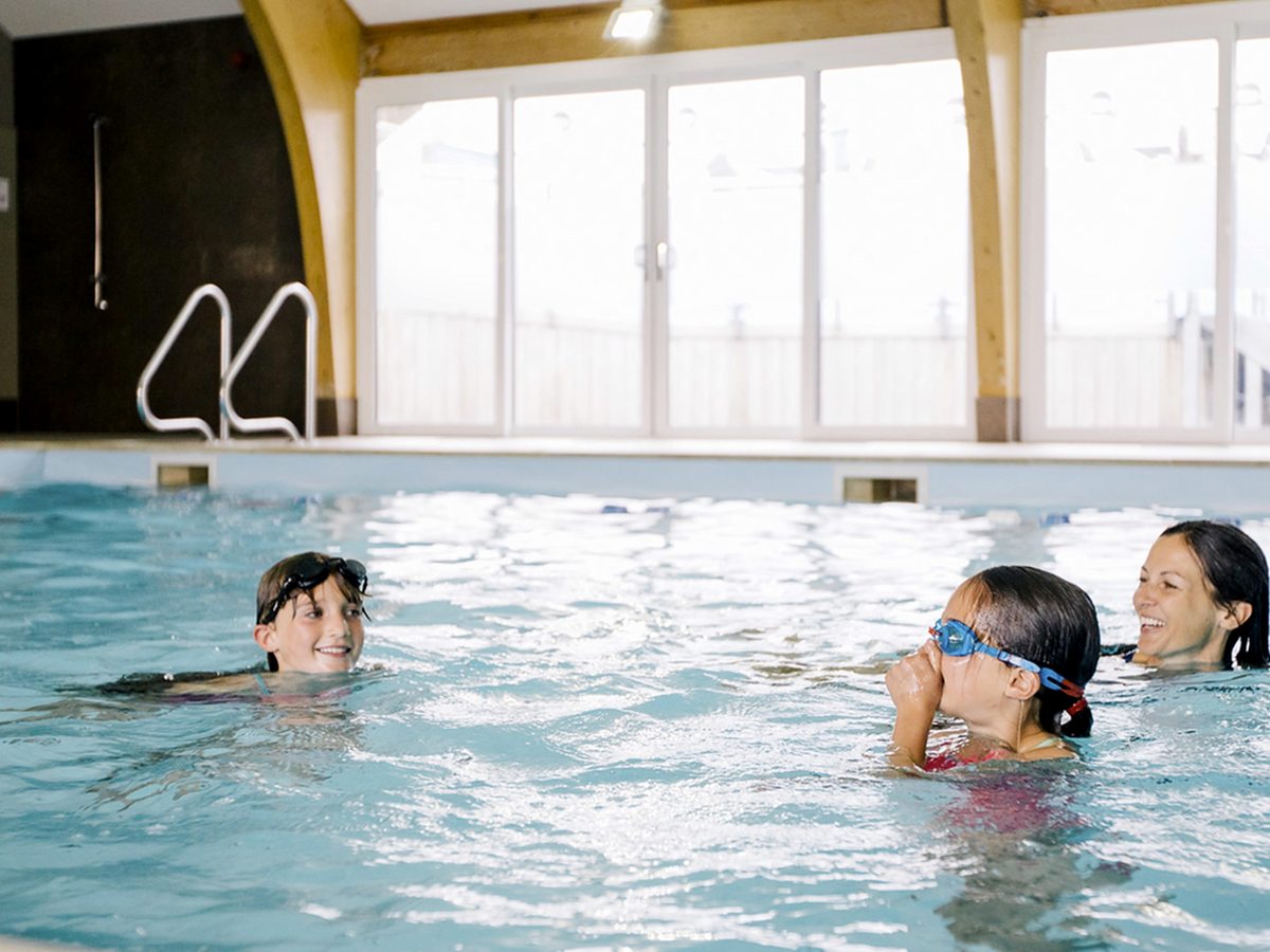 An indoor pool under a timber-arched roof with tall windows along one side, two children in goggles and an adult swimming in the pale blue water.