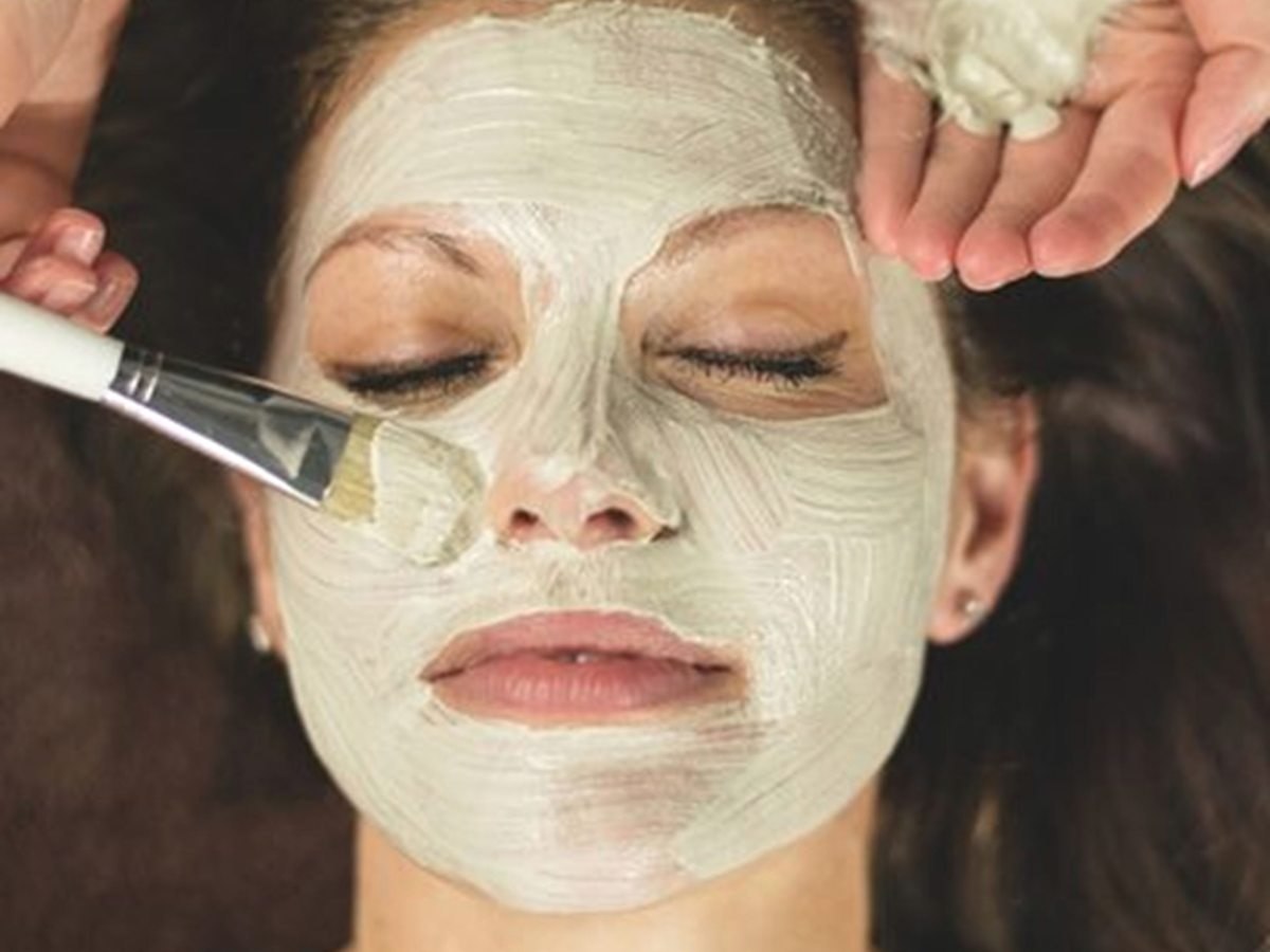 A therapist brushing a pale clay face mask onto a woman lying back with her eyes closed.