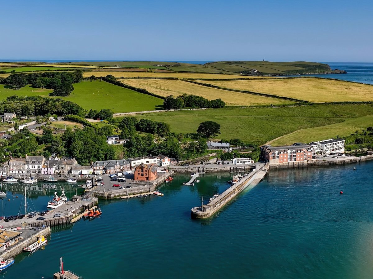 An aerial view of Padstow harbour with fishing boats and yachts against the quay, a stone breakwater, and patchwork fields running out to a headland.