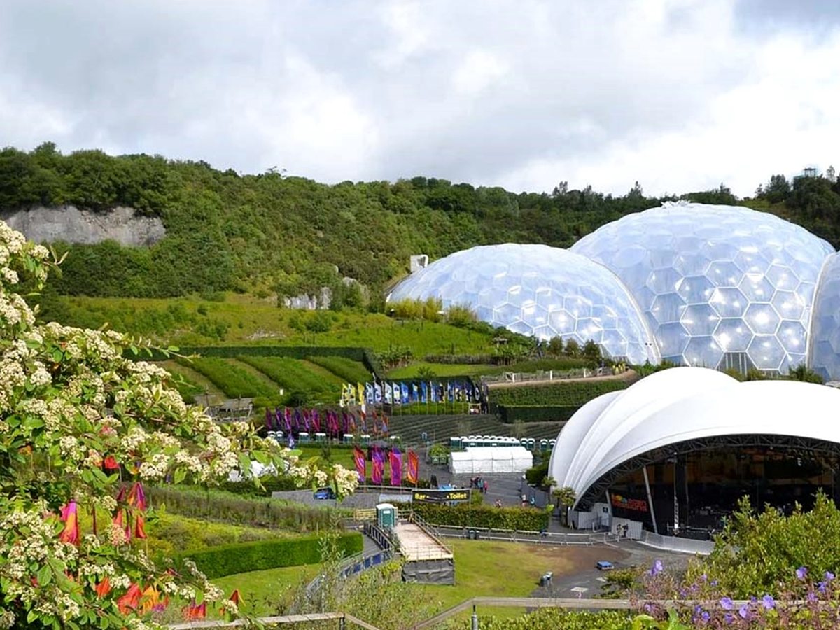 The white geodesic biome domes of the Eden Project set in a bowl of planted terraces and flower beds surrounded by wooded hillside.