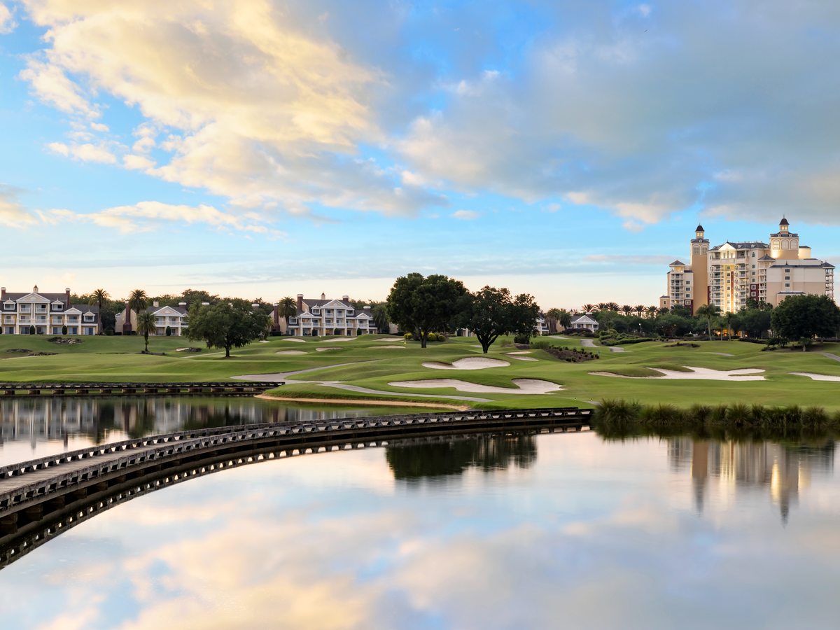 A golf course at dusk with a lake in the foreground, curved white footbridges crossing the water, fairways and low resort villas beyond under a pink sky.