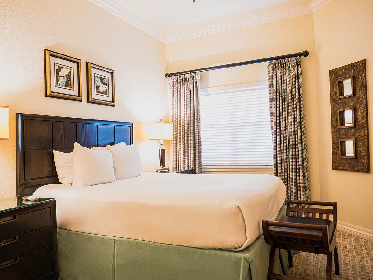 A bedroom with a king bed, a dark panelled headboard, a green valance, white bedding, twin bedside lamps and a curtained window.