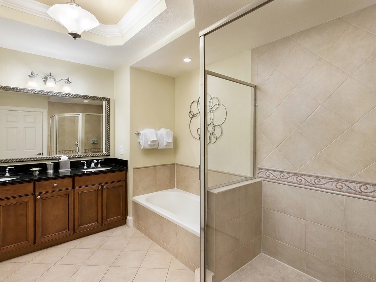 A large bathroom with a double wooden vanity and two washbasins, a wide mirror, an inset bath and a glass-enclosed walk-in shower, tiled throughout.
