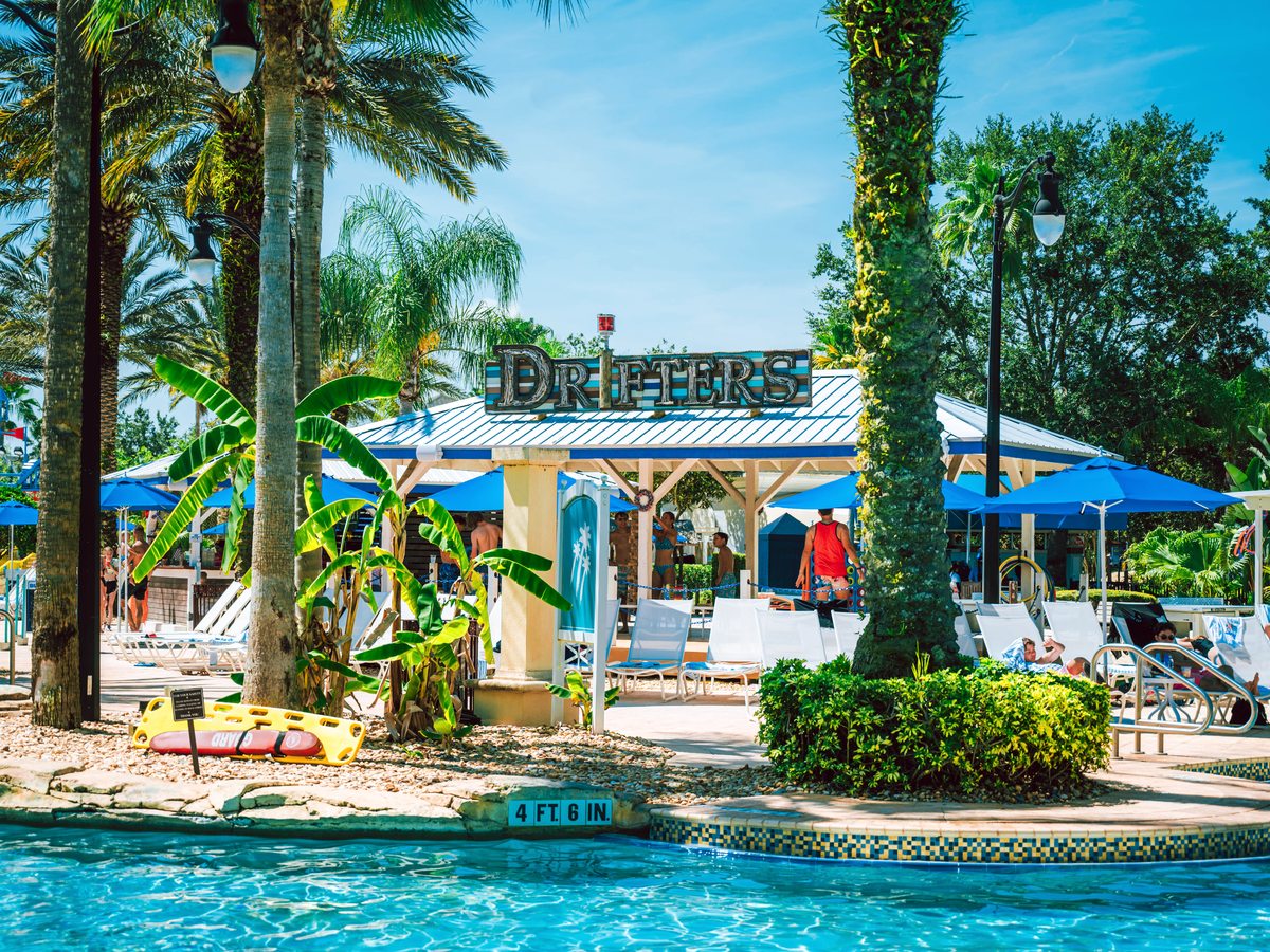 An open-air poolside bar building signed Drifters beside a swimming pool, with blue parasols, sun loungers and palm trees.
