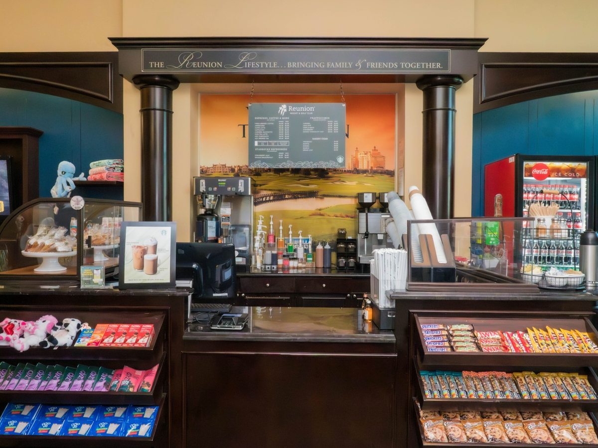 A grab-and-go cafe counter with an espresso machine, a digital menu overhead, chillers of bottled soft drinks and racks of packaged snacks and chocolate.