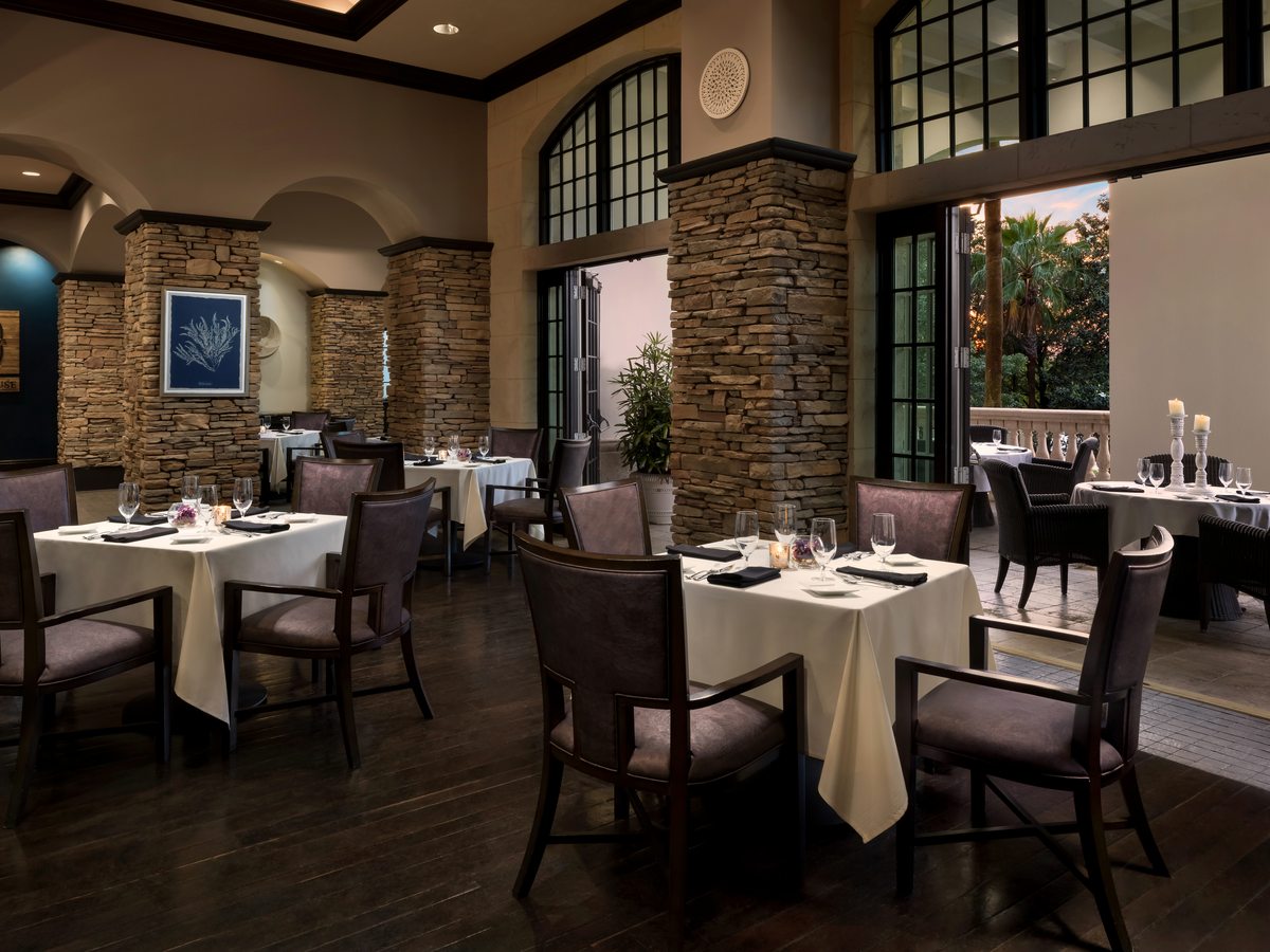 A formal restaurant dining room with white-clothed tables, dark upholstered chairs, stacked-stone columns, arched windows and a high ceiling.
