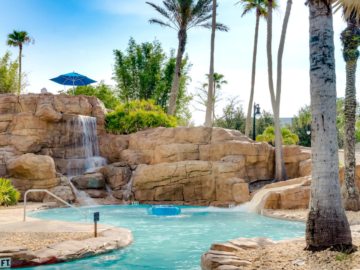 A turquoise pool channel running past an artificial rock formation with a waterfall, palm trees and tropical planting, a lifebuoy and a directional signpost at the water's edge.