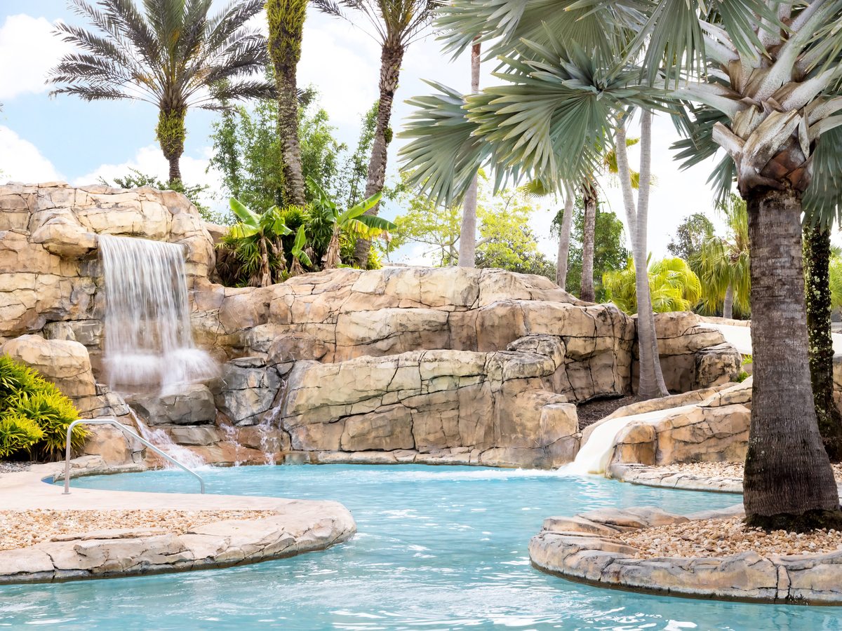 Two waterfalls tumbling over a curved artificial rock formation into turquoise water, with handrails at the pool entry and palm trees behind.