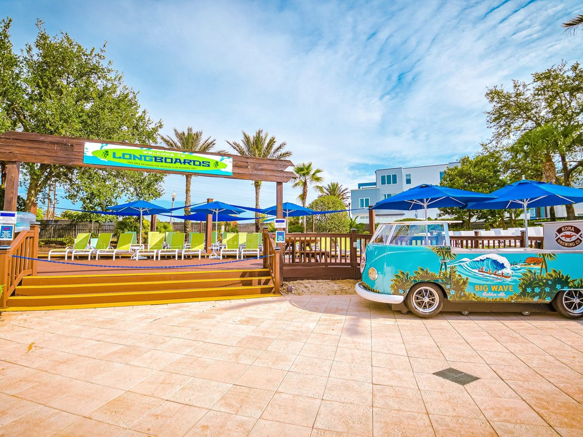 A poolside food outlet built around a vintage-style van with surf graphics on a wooden deck, with blue parasols, shaded tables and lime-green loungers.