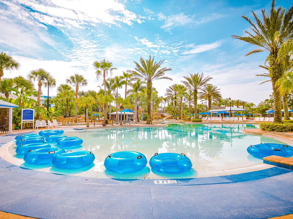 A shallow lazy river with blue inflatable tubes stacked at the entry point, palm trees and a pool deck with sun loungers and shade structures.