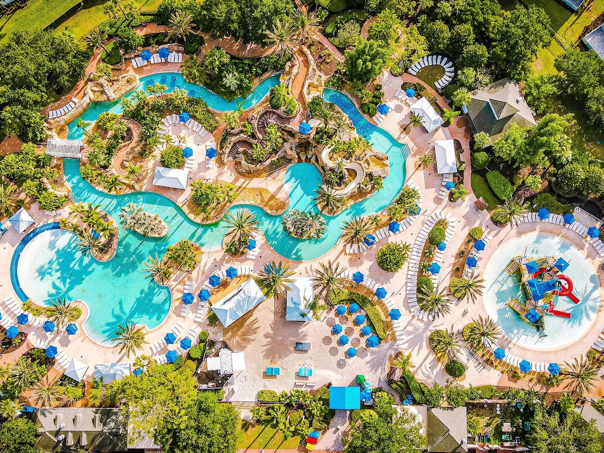 An overhead aerial of the water park showing a winding lazy river and interconnected pools threading between landscaped islands, sun loungers and parasols all around.