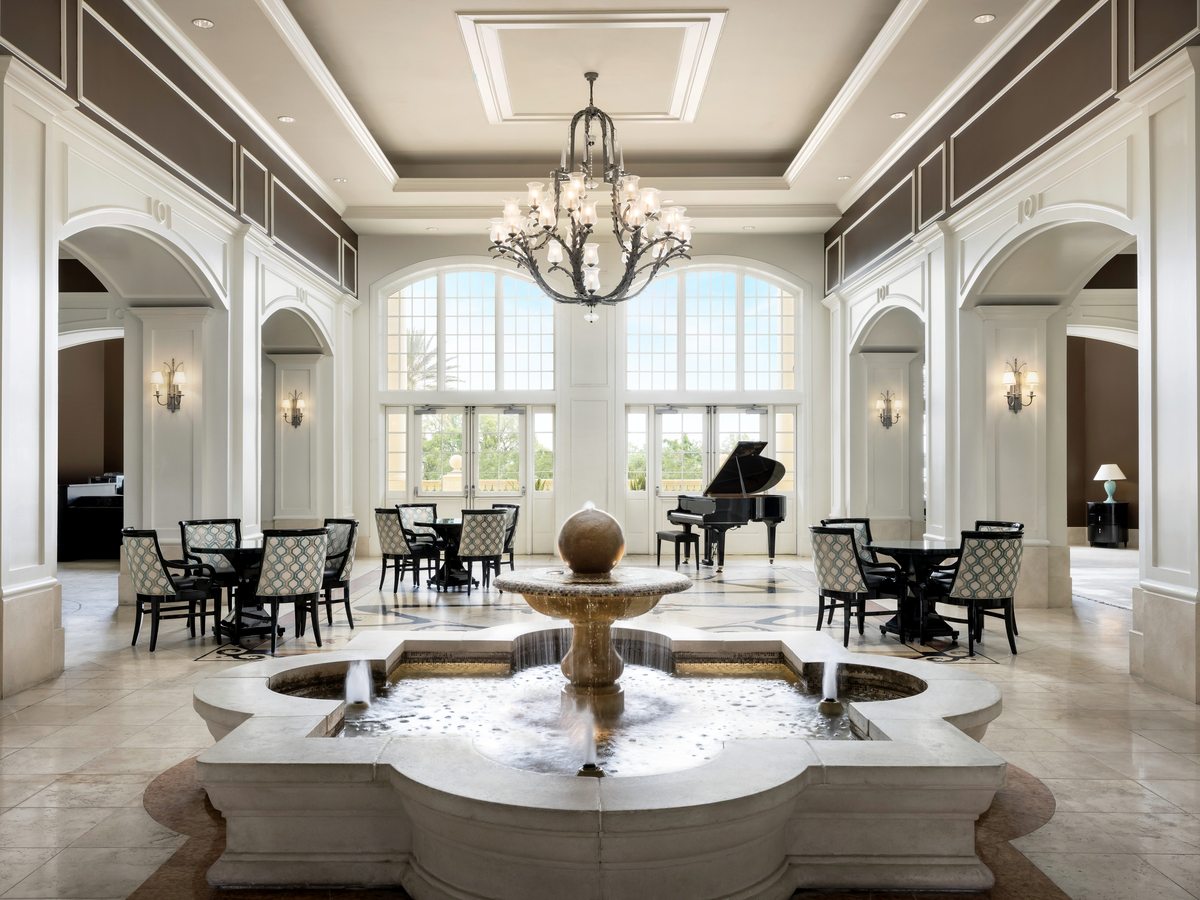 A double-height lobby with a marble floor, a tiered stone fountain at its centre, chandeliers, tall arched openings and seating around the edges.