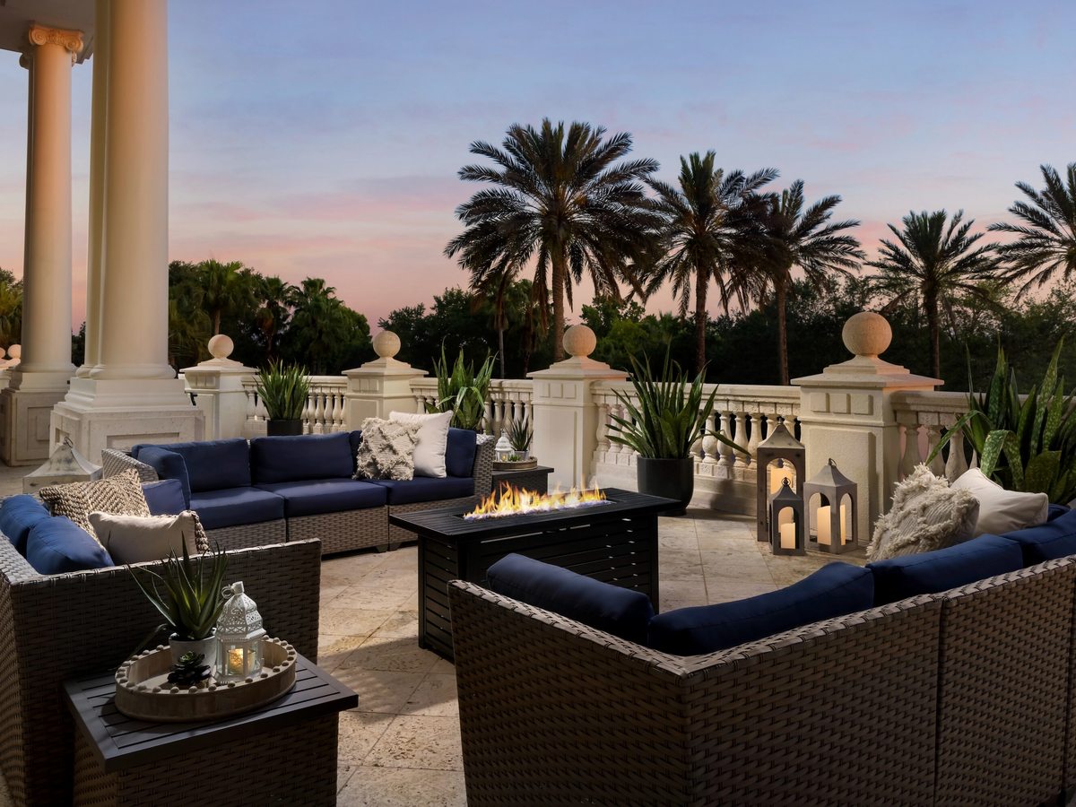 A rooftop terrace at sunset with wicker sofas and armchairs, blue cushions, low tables, lanterns and a classical balustrade looking over palm trees.
