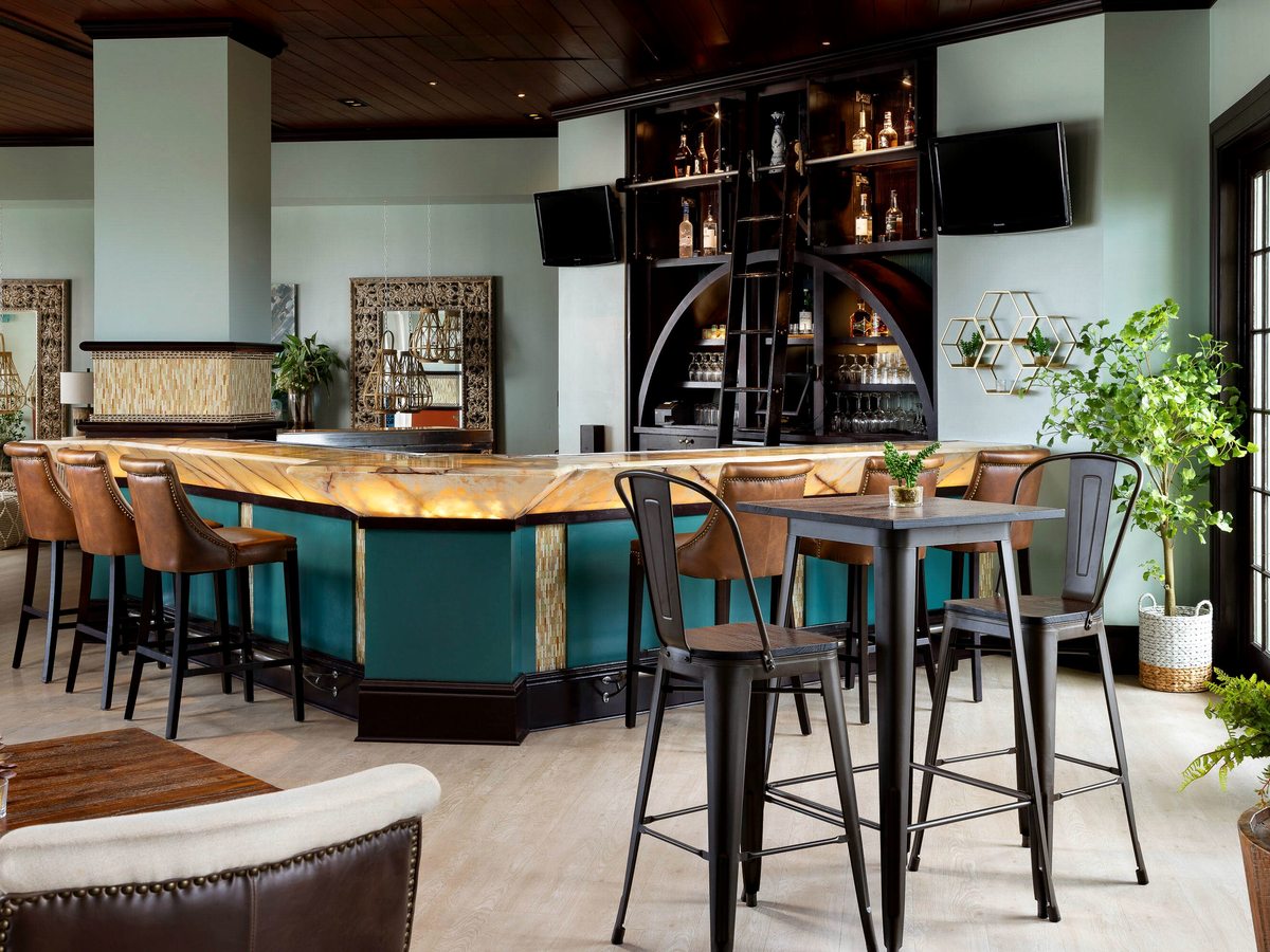 An indoor bar with a turquoise-panelled counter, leather stools, a wall of bottles reached by a rolling library ladder, two televisions and lounge seating.
