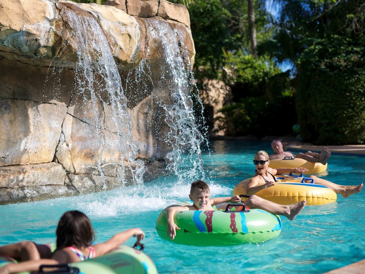 People floating on inflatable tubes along a lazy river beneath a rock waterfall, with palm trees and tropical planting around the turquoise water.