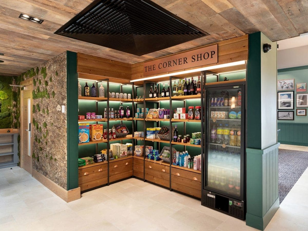 A compact wood-fronted shop under a sign reading The Corner Shop, with shelves of cereal, snacks, drinks, wine and toiletries beside a glass-fronted chiller.