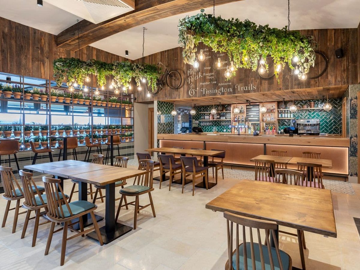 A rustic restaurant dining room with wooden tables and chairs, a long bar counter with a green tiled splashback, timber-clad walls and troughs of hanging greenery lit by pendant bulbs.