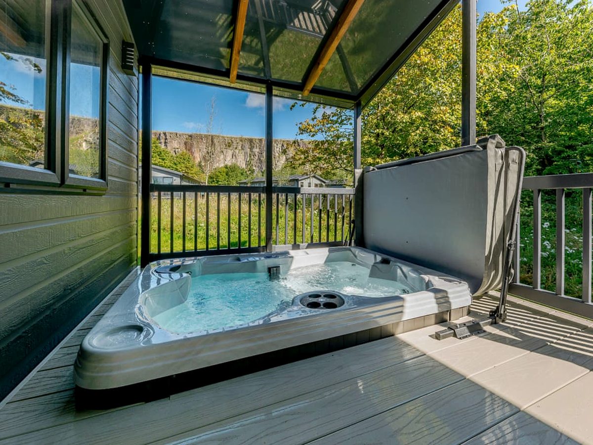 A bubbling hot tub on a sheltered timber deck beside a lodge's grey cladding, with deck railings, trees, neighbouring lodges and a high rocky quarry face beyond.