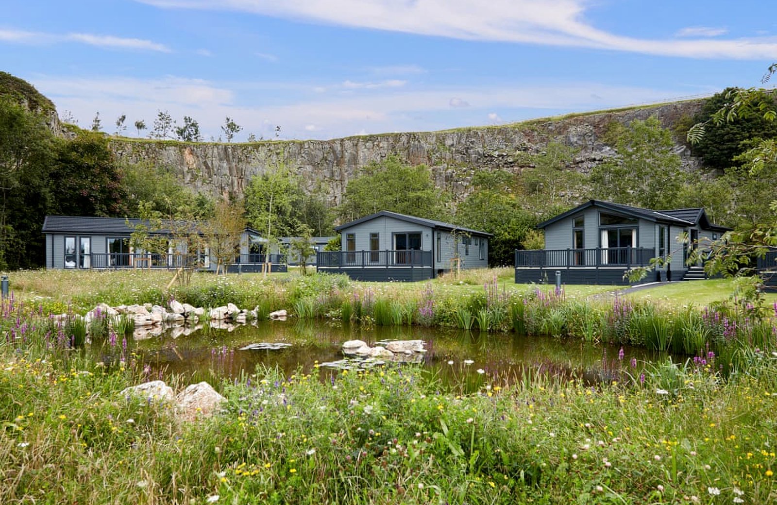 Rivendale Lodge Retreat: timber lodges beyond a wildlife pond and wildflower meadow, with the limestone quarry face rising behind, inside the Peak District National Park.