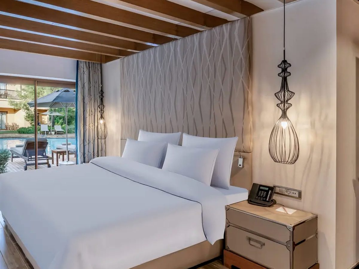 A double room with a timber-beamed ceiling and pendant lamps, its sliding glass doors open onto a decked terrace with loungers and a parasol beside one of the resort's pools.