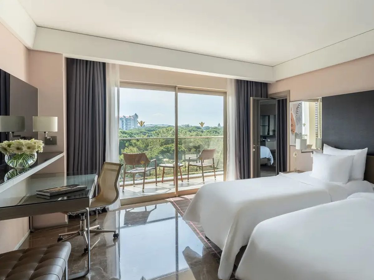 A twin room with two single beds, a desk and chair, a glossy tiled floor and sliding doors onto a furnished balcony above the gardens.