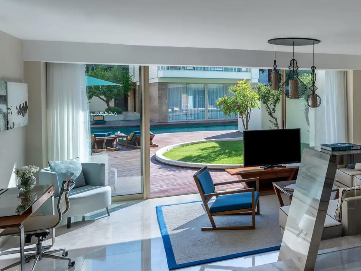 A suite living area with a blue rug, armchairs, a desk and a television, its sliding doors open to a lawn and a pool running between the accommodation blocks.