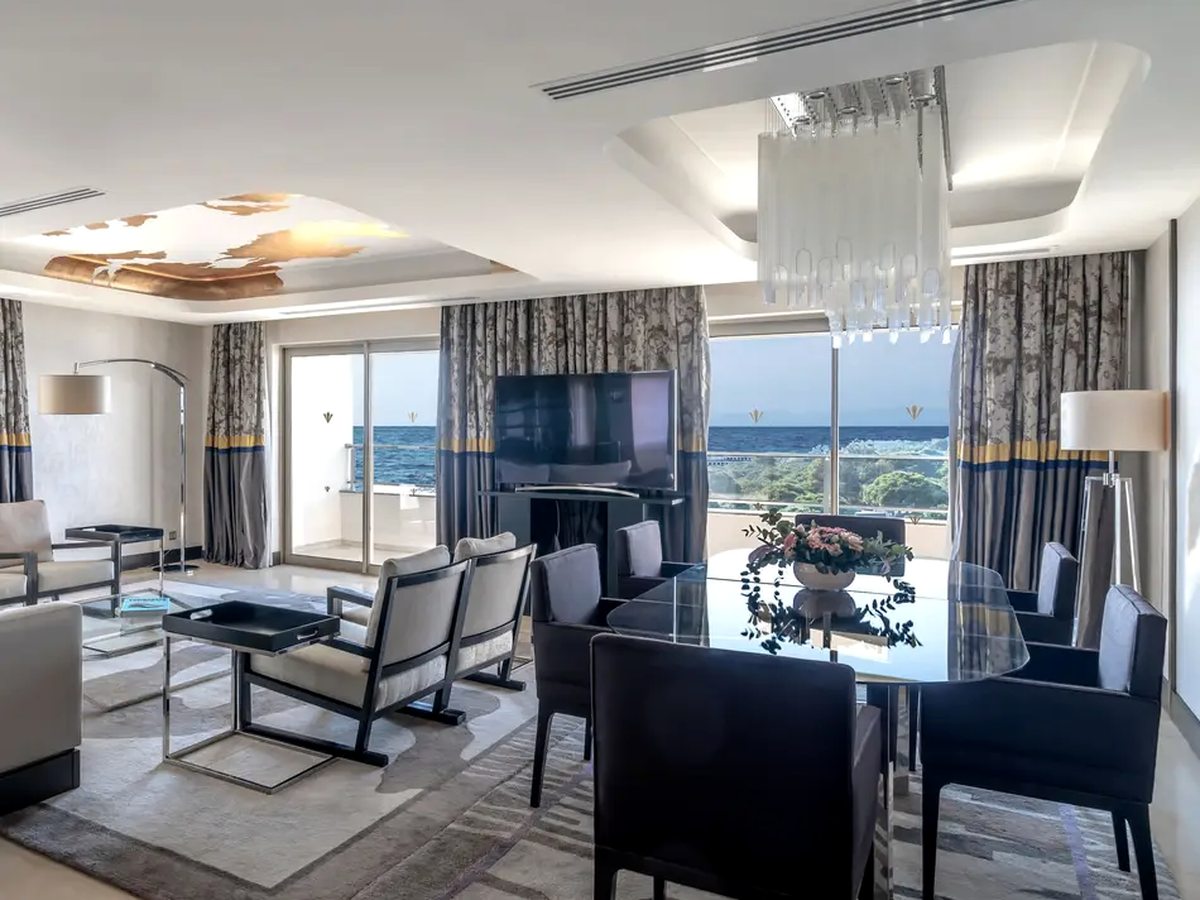 A large suite room with a dining table and chairs at one end, a lounge area and chandelier at the other, and full-height doors to a balcony above the trees.