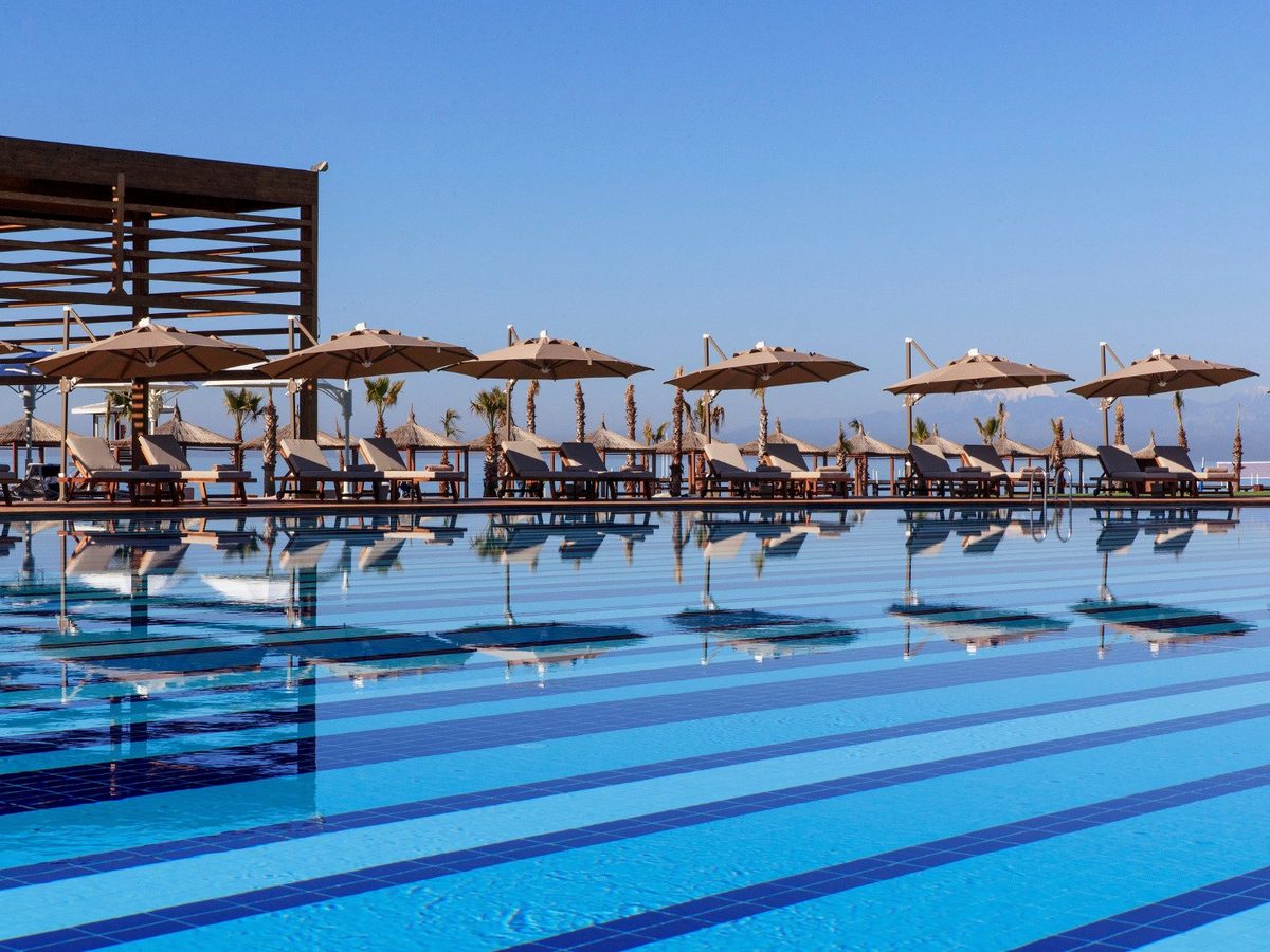A wide pool with broad blue stripes across its floor, a row of parasols and wooden loungers along the far edge and mountains on the horizon.