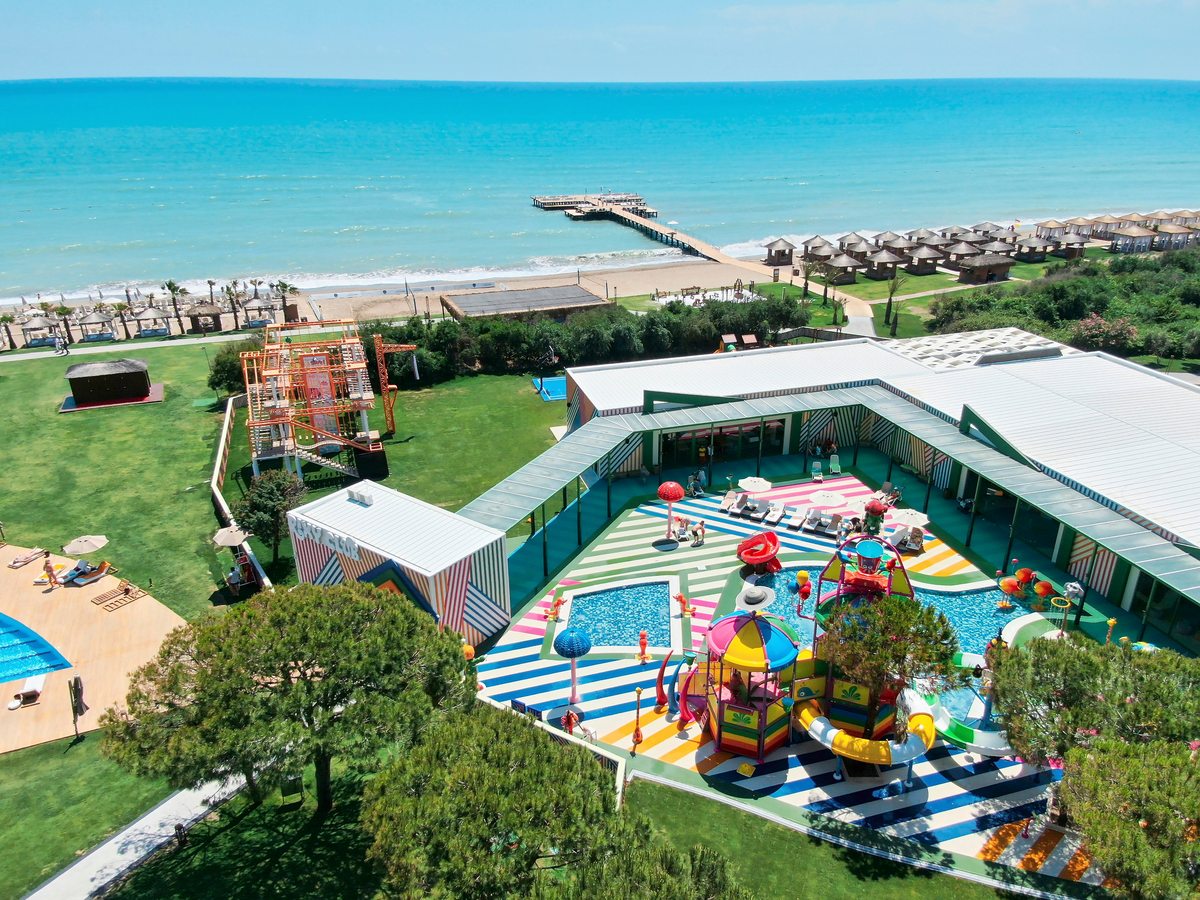 A brightly striped children's water-play area with slides, tipping buckets, spray features and shallow pools, on a lawn behind the beach.
